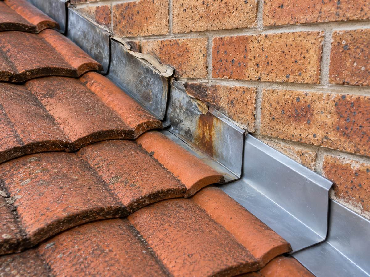 Metal step flashing where a tile roof meets a brick wall, with lifted sealant and a rust stain beside a sound new section. Illustration