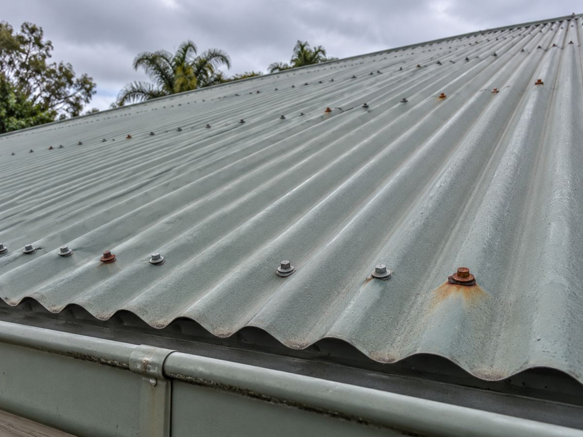 A grey-green corrugated metal roof with rows of screws, a few rusted heads and a rust patch around one fastener above the gutter. Illustration